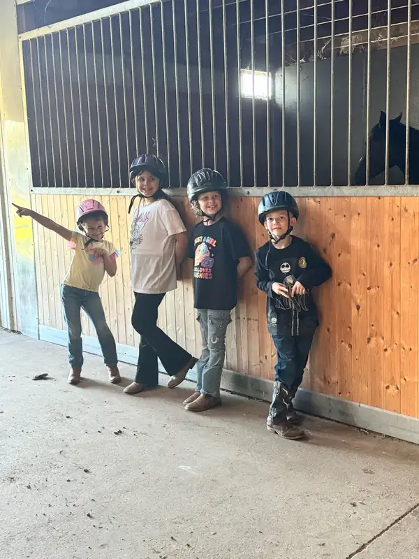 Four children in riding helmets standing against a stall wall in the barn