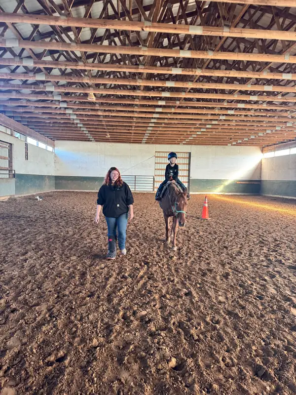 An instructor leading a young rider in a helmet on a pony across the indoor arena