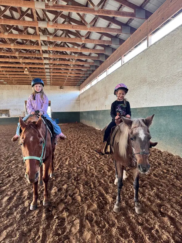 Two young riders in helmets on horses in the indoor arena