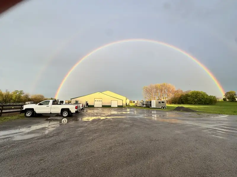 A full rainbow arching over the main barn and a parked white pickup truck